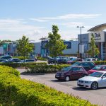 Retail park on a sunny day with parked cars, featuring stores like DW Sports and Fitness, Next Home, and Boots