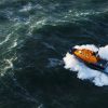 Orange lifeboat navigates choppy green sea, creating white waves as it moves swiftly across the water