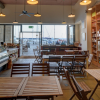 Cosy cafe interior with wooden tables, deli counter, and shelves of beverages, overlooking marina through large windows