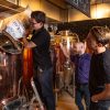 Brewers adding ingredients to a copper brewing tank in a brewery, surrounded by equipment and observing brewing process