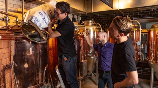 Brewers adding ingredients to a copper brewing tank in a brewery, surrounded by equipment and observing brewing process