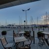 Outdoor café with wooden tables and chairs overlooking a marina with yachts and a clear blue sky, ideal for relaxation