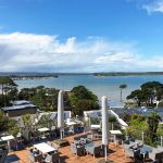 Scenic coastal view with outdoor terrace seating, overlooking a bay and boats under a bright blue sky