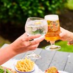 Friends toasting with beer and gin at an outdoor picnic, food and greenery in the background