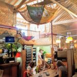 Cosy café interior with colourful decor, wooden beams, and customers enjoying drinks at a communal table in rustic setting