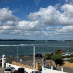 Scenic view of harbour with parked cars, jet skis on water, cloudy sky, and distant islands.
