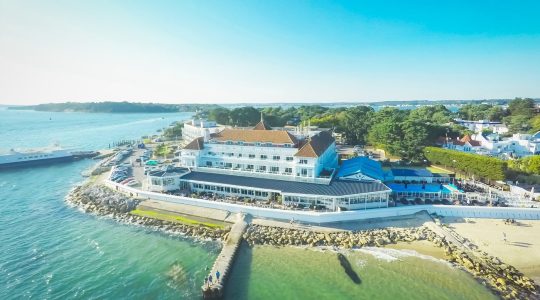 Aerial view of a seaside hotel with a sandy beach and vibrant turquoise water, surrounded by lush greenery