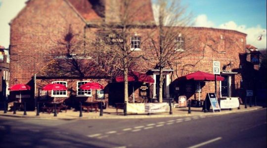 Historic brick pub with red umbrellas on a sunny street in the UK, offering charming outdoor seating