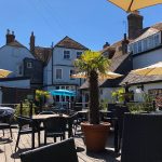 Sunny outdoor cafe with black wicker tables and chairs, under cream umbrellas, surrounded by quaint historic buildings