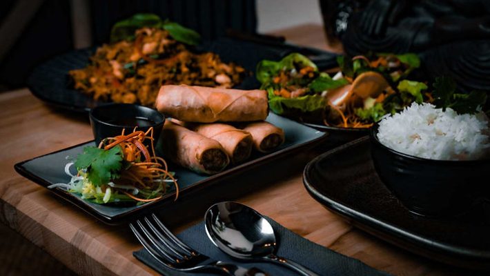 Asian food platter with spring rolls, rice, and salad on a wooden table, cutlery placed nearby