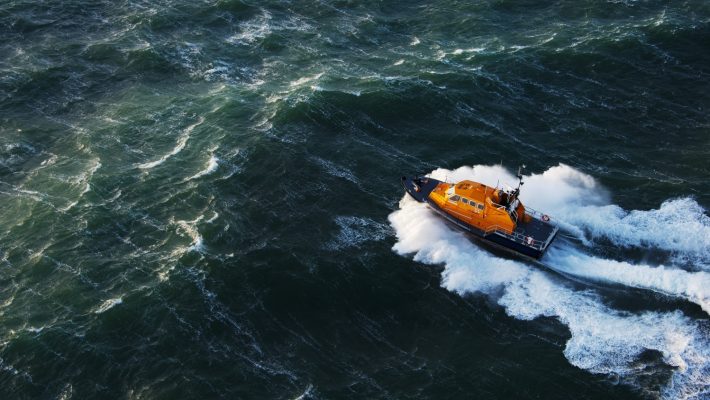 Orange lifeboat navigates choppy green sea, creating white waves as it moves swiftly across the water