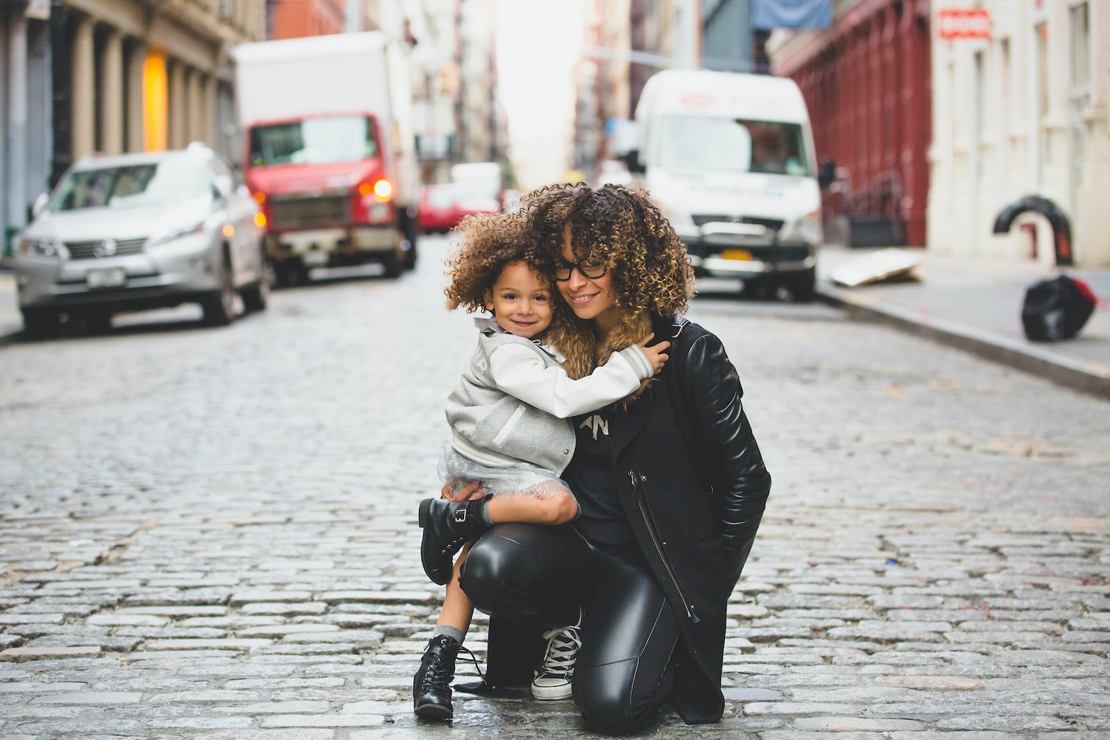 Mother and daughter hugging on a cobblestone street, surrounded by urban traffic and buildings