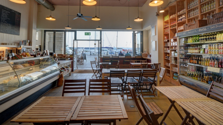 Cosy cafe interior with wooden tables, deli counter, and shelves of beverages, overlooking marina through large windows