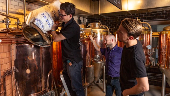 Brewers adding ingredients to a copper brewing tank in a brewery, surrounded by equipment and observing brewing process
