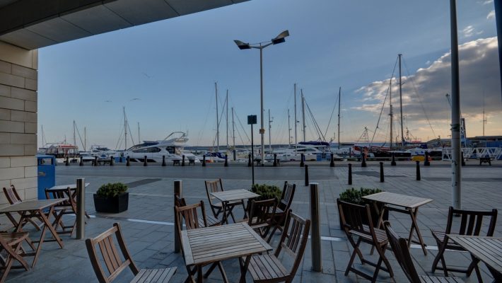 Outdoor café with wooden tables and chairs overlooking a marina with yachts and a clear blue sky, ideal for relaxation