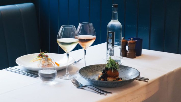Elegant restaurant table setting with gourmet dishes, white and rosé wine glasses, and water bottle on a white tablecloth
