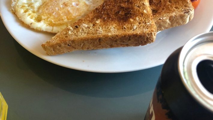 Full English breakfast with toast, egg, beans, sausage, butter, and a soft drink can on a glass table