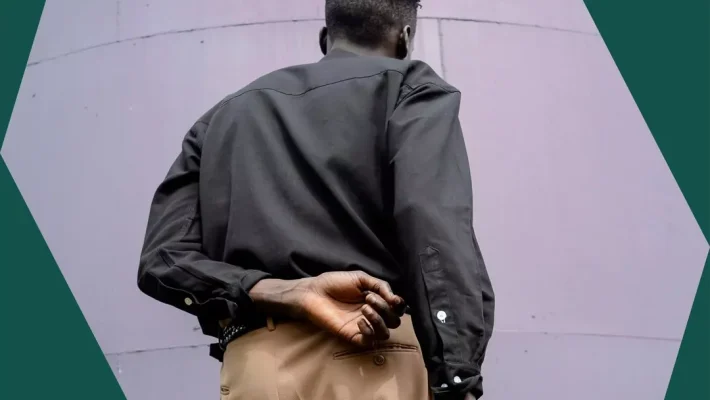 Man in a black shirt and tan trousers, standing against a purple wall, with his hand behind his back