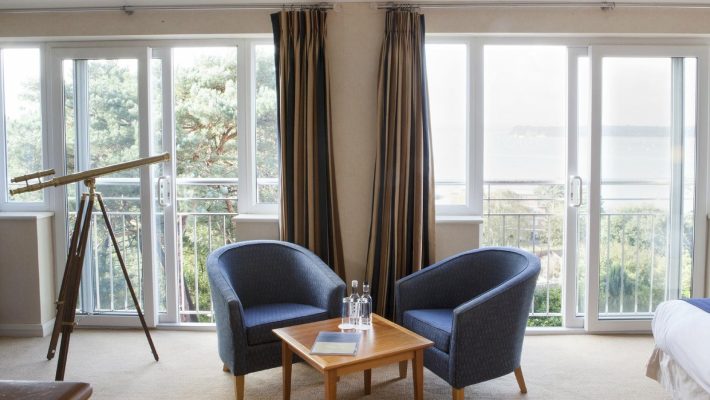 Hotel room with sea view, featuring two chairs, table, telescope, large windows, and curtains on beige carpet