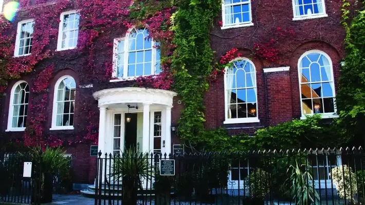 Historic red-brick building with ivy-covered facade and arched windows, framed by a black wrought-iron fence
