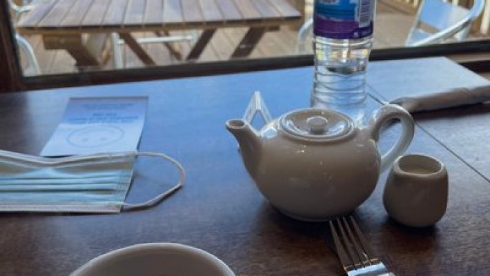 Tea set on a café table with a view of the outdoor seating area through the window, including water bottle and mask