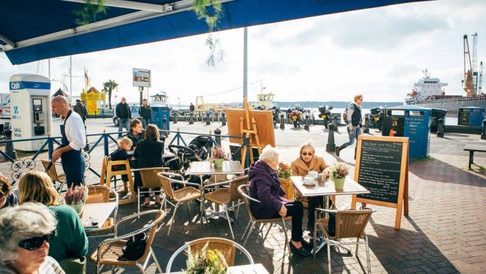 People dining at a waterfront cafe with a view of ships and a vibrant outdoor atmosphere