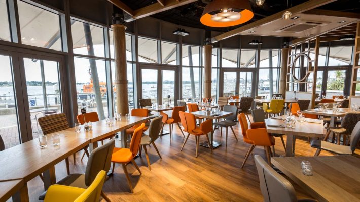 Modern waterfront restaurant interior with wooden flooring and colourful chairs, offering a view of the harbour
