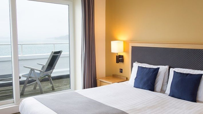 Modern hotel room with ocean view, featuring double bed, pillows, and balcony with deck chair and glass doors