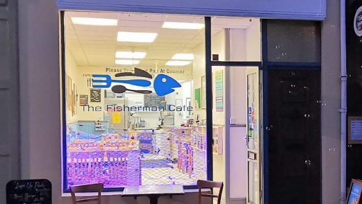 Front view of The Fisherman Café with a cosy seating area outside and glowing interior lighting