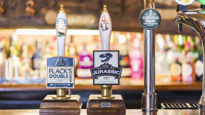 Pub bar with Flack Manor and Jurassic beer taps, including Curious Brew tap handle in a lively background setting