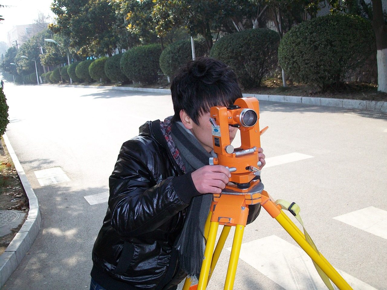 Person using a theodolite for surveying on a sunny day, wearing a black jacket and scarf