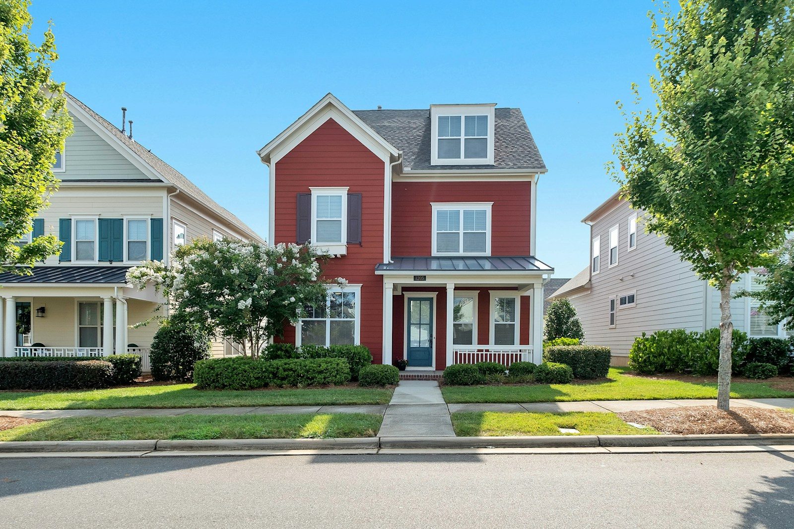 Charming red two-storey house with white trim and front porch surrounded by greenery under a clear blue sky